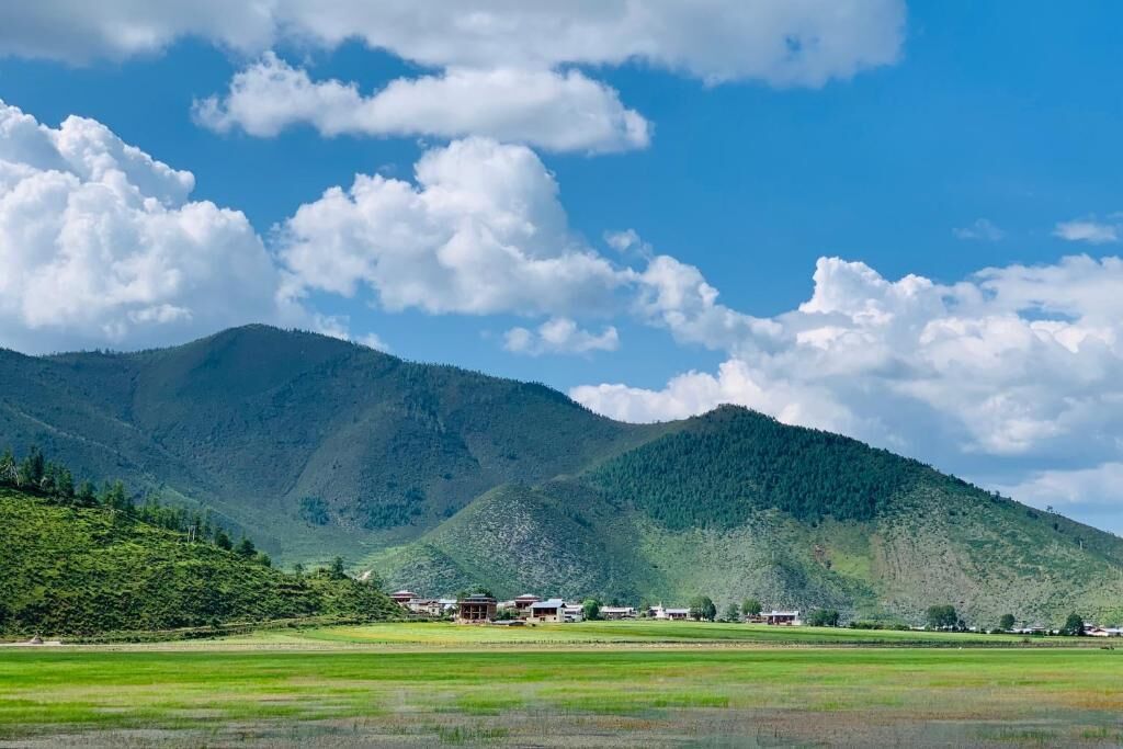 Un paisaje montañoso con cielo despejado y nubes dispersas. Se observan edificios con tejados rojos en primer plano, probablemente parte de un pueblo o pequeña ciudad.
