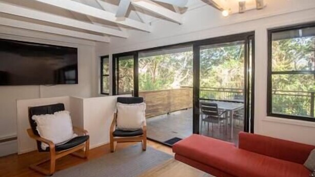 A modern living room with a red sofa, two wooden chairs, a TV, and large windows offering a view of the outdoors. - 20 Yarrong Rd - a very large house split over three levels (Point Lookout)