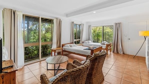 A room with two beds, a small table, wicker furniture, and large windows with curtains.