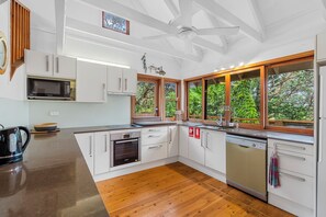 A modern kitchen with white cabinets, a microwave, oven, and dishwasher. There are large windows with a view of greenery outside. - 20 Yarrong Rd - a very large house split over three levels (Point Lookout)