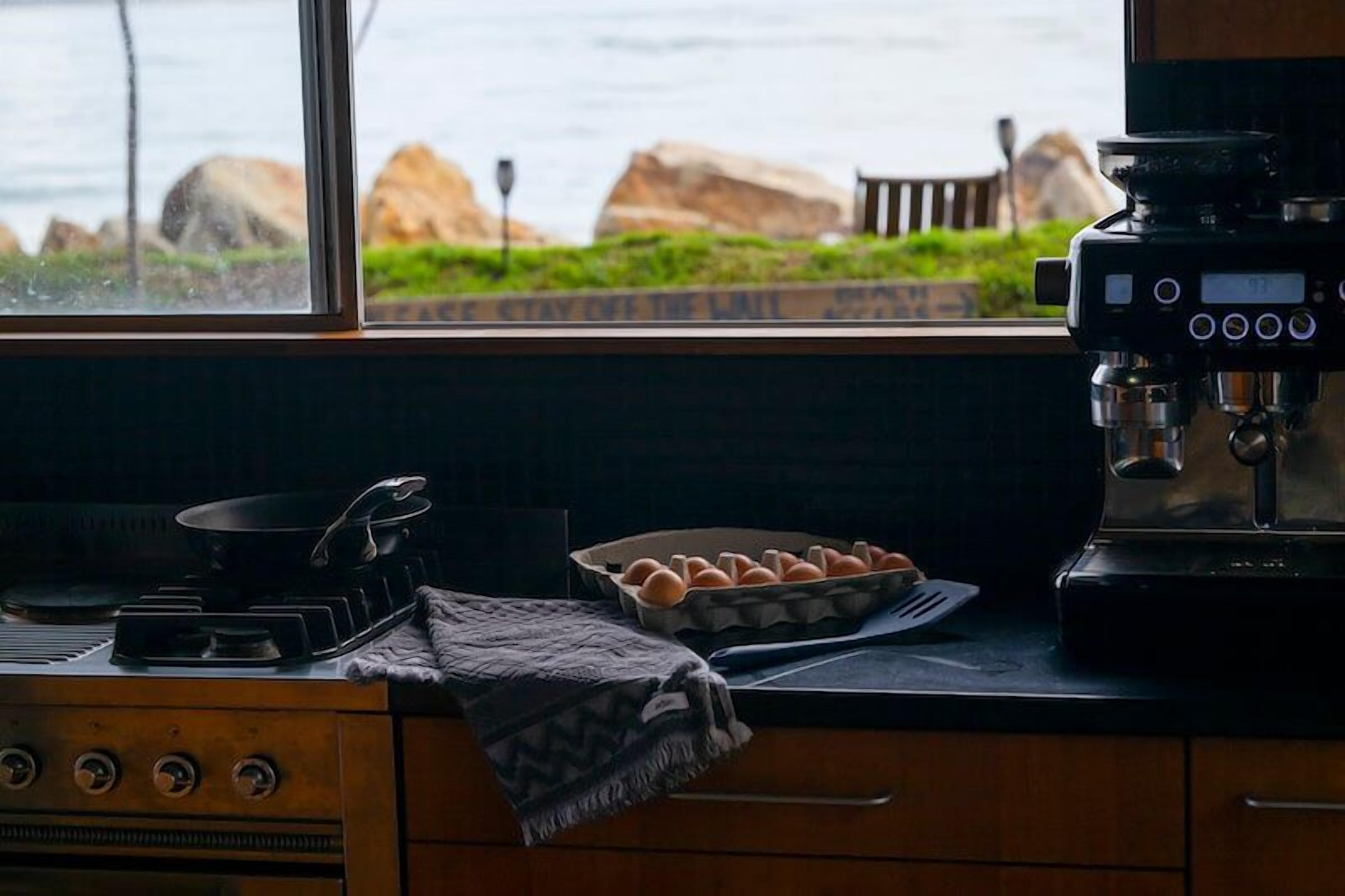 A kitchen with a stove, a coffee maker, and a view of the ocean through a window.