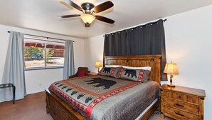 A bedroom with a wooden bed, a ceiling fan, a window with curtains, a nightstand with a lamp, and a view of the outside.