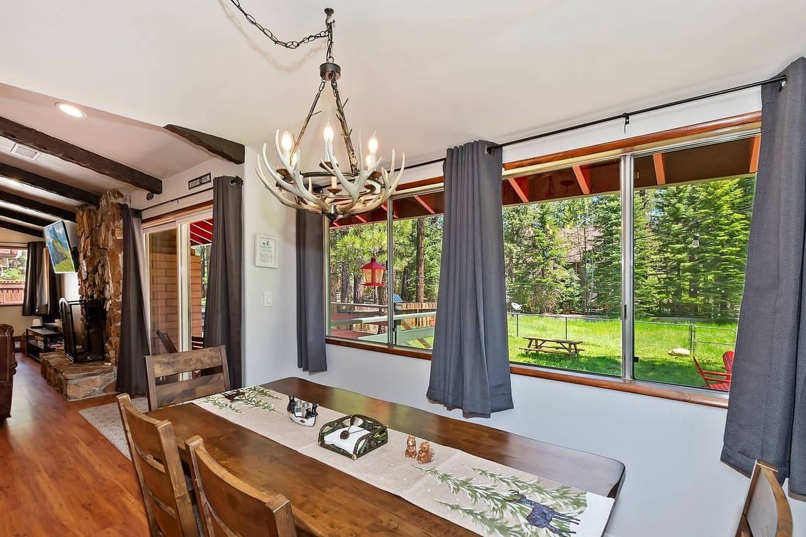 A dining area with a long table, chairs, a chandelier, and large windows with curtains.