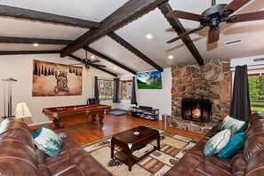 A spacious living room with a stone fireplace, a pool table, leather sofas, and a ceiling fan.