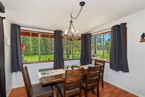 Comedor con una mesa dispuesta para cuatro, una araña y ventanas grandes con cortinas.