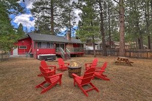 A rustic outdoor seating area with red Adirondack chairs and a fire pit, surrounded by a wooden fence and tall trees.