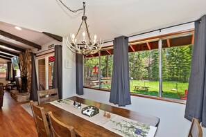 Un comedor con mesa de madera, lámpara de araña y ventanas amplias con vistas a un jardín.