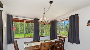 A dining area with a wooden table, four chairs, a chandelier, and large windows with curtains.