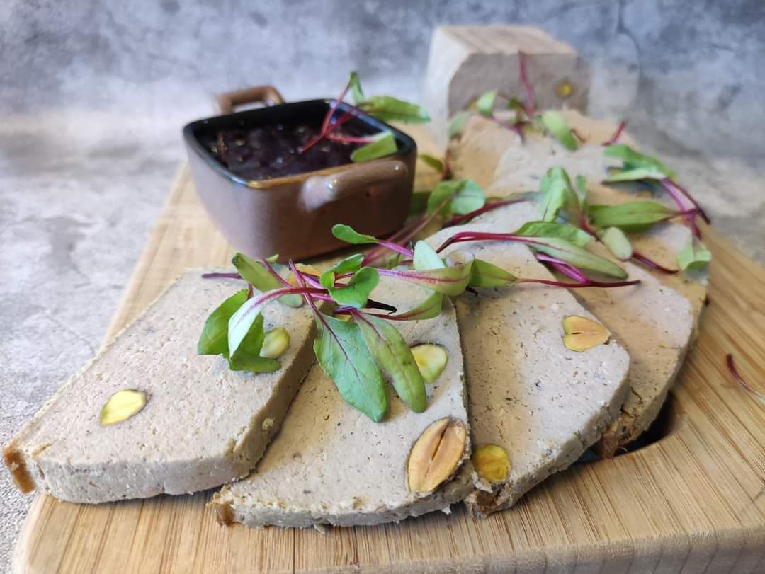 Una tabla de madera con rebanadas de paté adornadas con hierbas y nueces, y un cuenco pequeño de chutney.