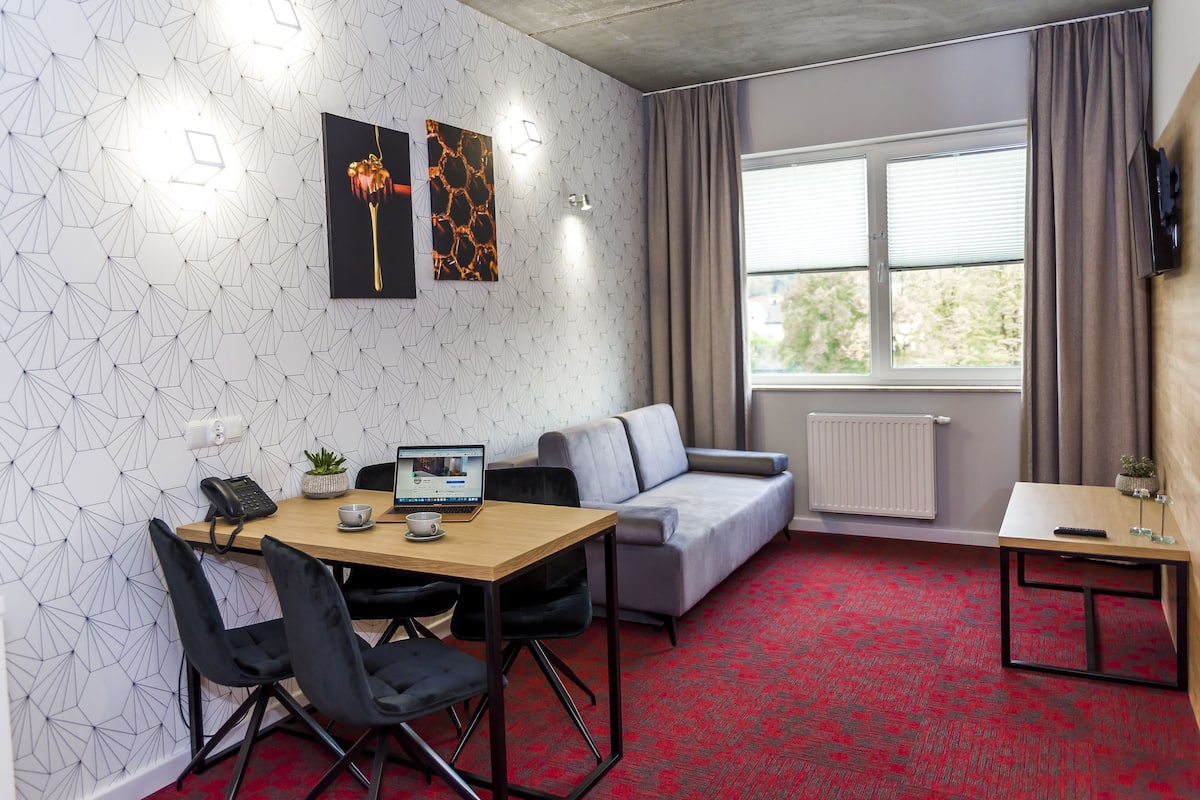 A modern room with a geometric-patterned wall, a grey sofa, a wooden table with a laptop, and a red carpet.