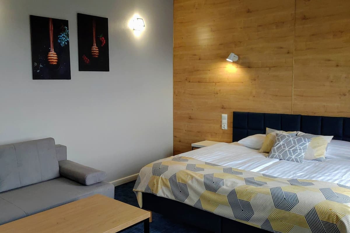 A room with a bed, a sofa, and a wooden headboard. There are wall-mounted lights and a wall-mounted artwork.