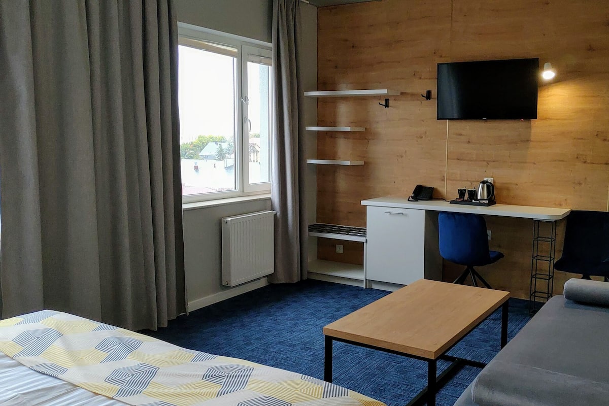 A small room with a bed, a desk, a chair, a TV mounted on the wall, and a window with curtains.