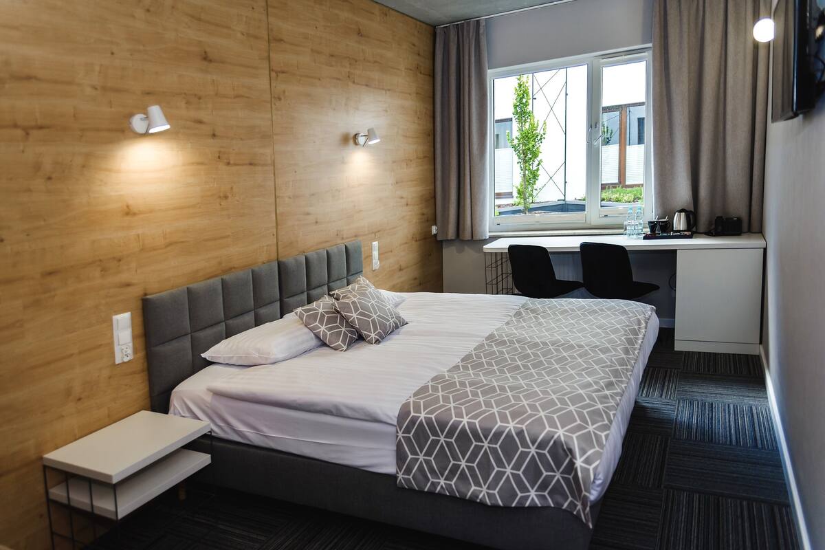 A hotel room with a bed, bedside table, desk, and a window with curtains.
