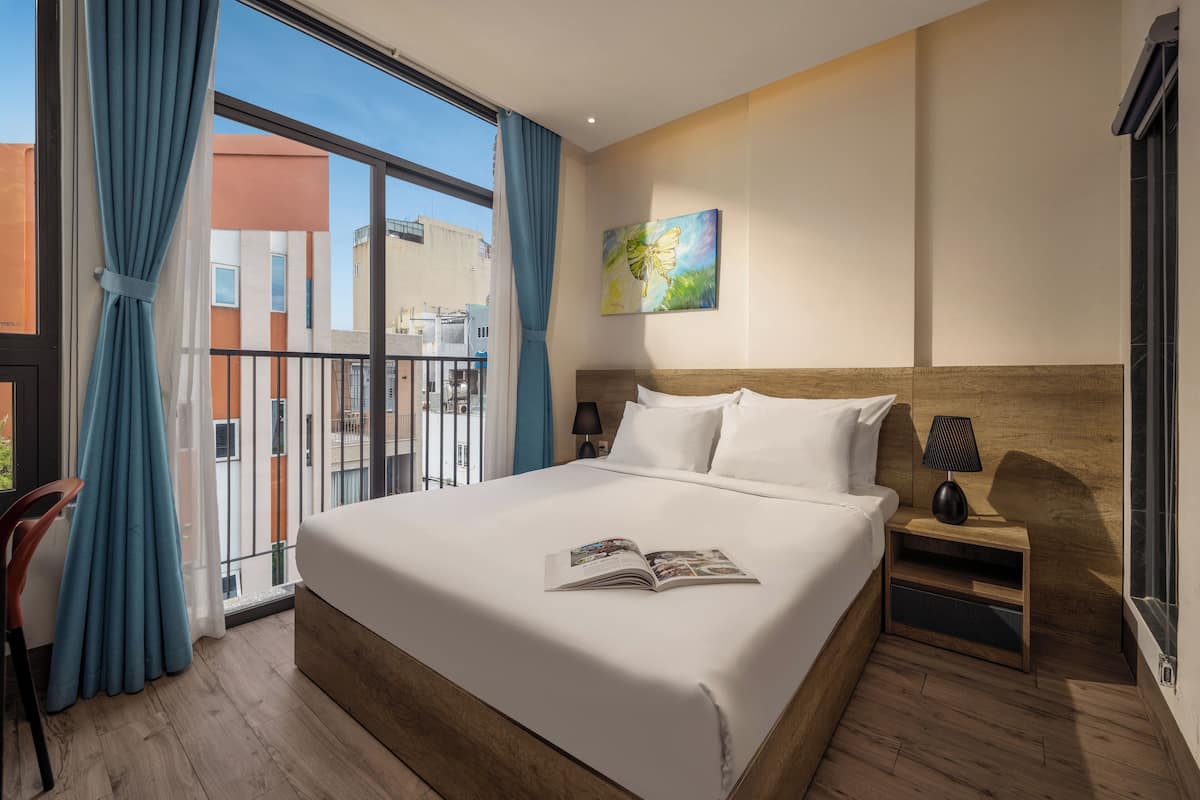Ein Hotelzimmer mit einem großen Bett, einem hölzernen Kopfteil, einem Nachttisch mit Lampe, einem Fenster mit blauen Vorhängen und einem Bild an der Wand.