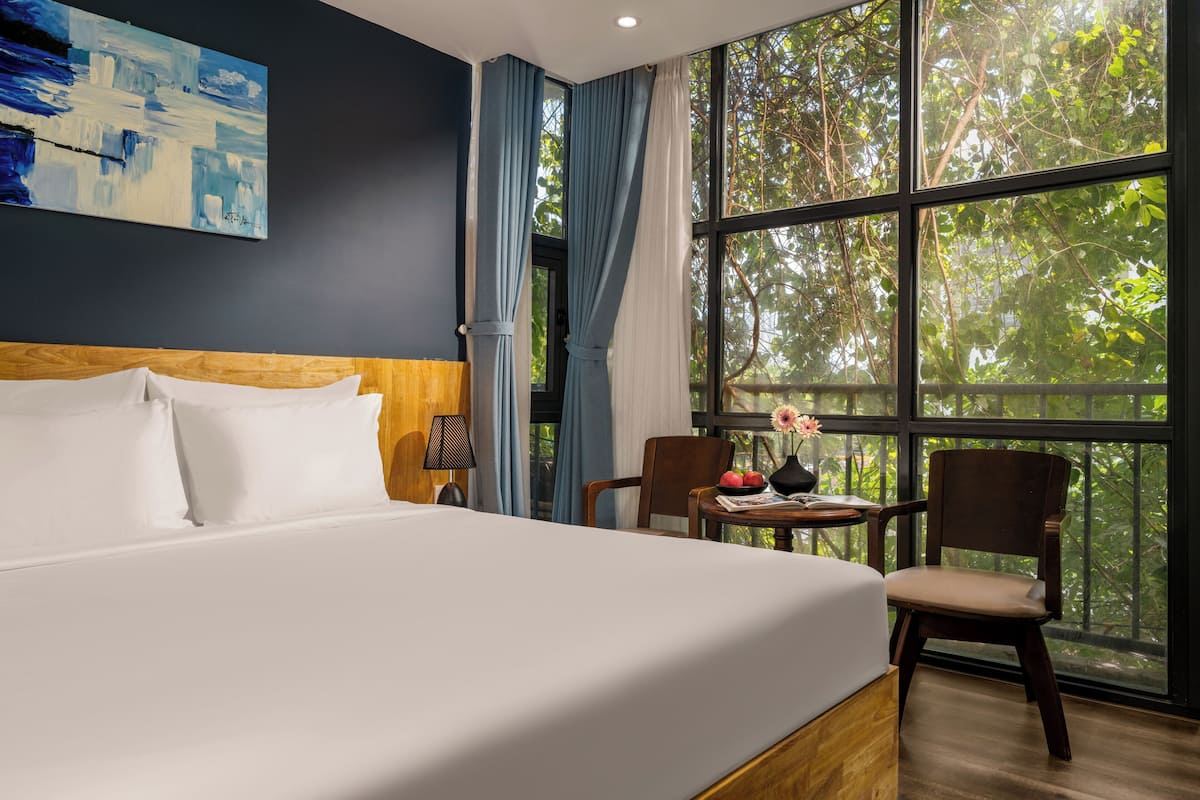 Ein Hotelzimmer mit einem großen Bett, einem Holzkopfteil, einem Schreibtisch mit Stuhl und Blick auf Bäume durch große Fenster.