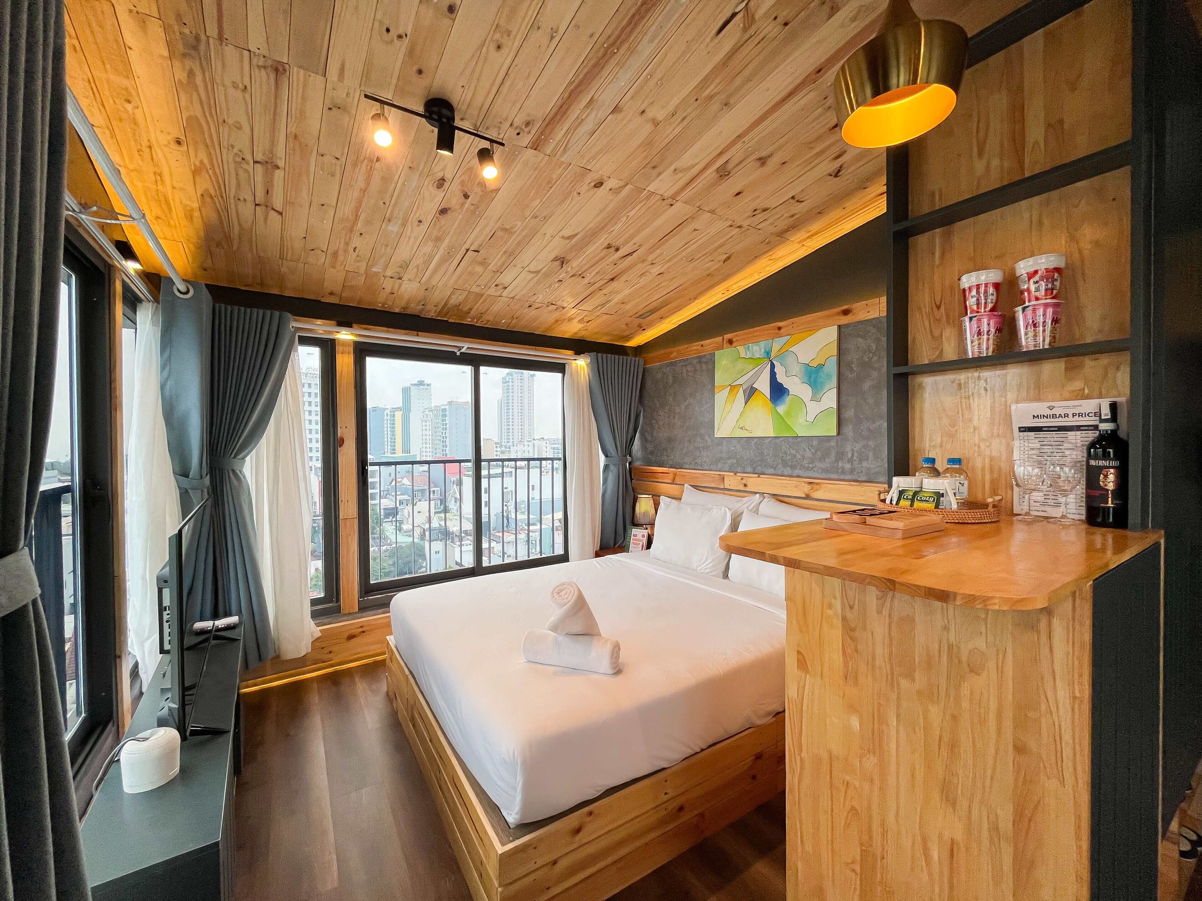 Ein Hotelzimmer mit einem großen Bett, Holzboden, Blick auf die Stadt, einer Minibar und einem Bild an der Wand.