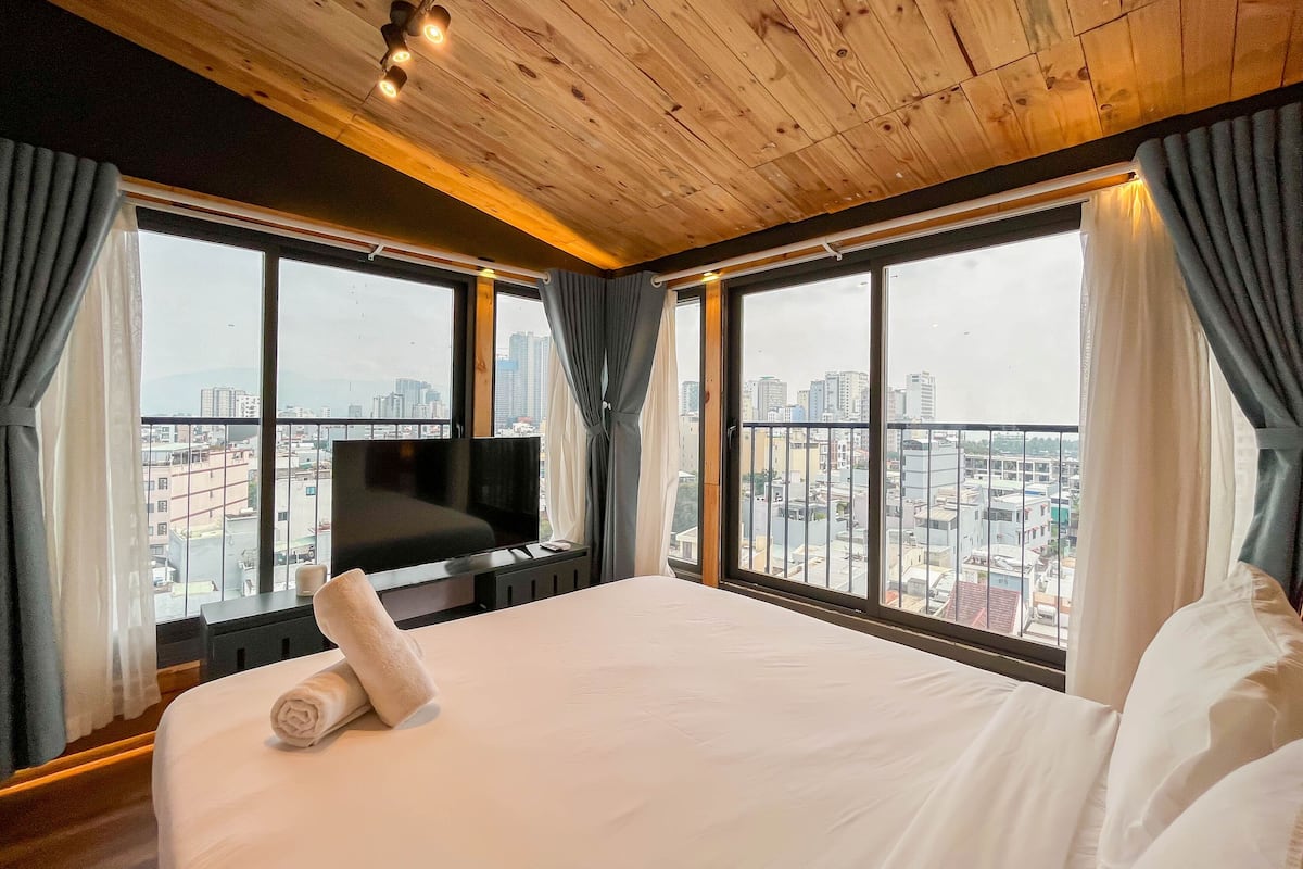 Ein Hotelzimmer mit einem großen Bett, einem Flachbildfernseher und Blick auf die Stadt durch große Fenster.