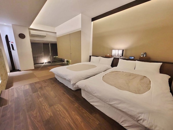 A hotel room with two beds, a wooden floor, a large window, and a bathroom visible in the background.