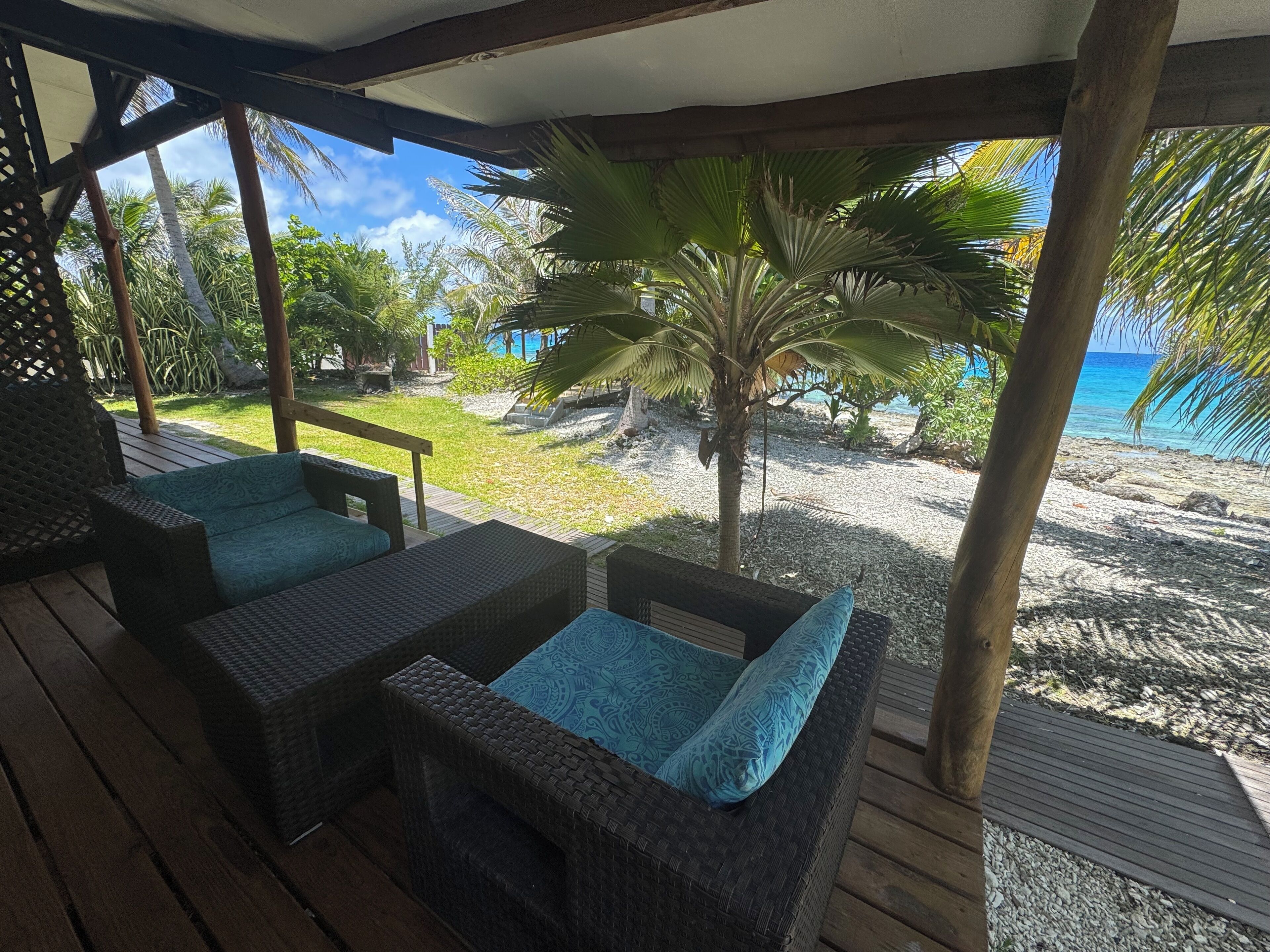 A covered outdoor seating area with wicker furniture and a view of a beach and palm trees.