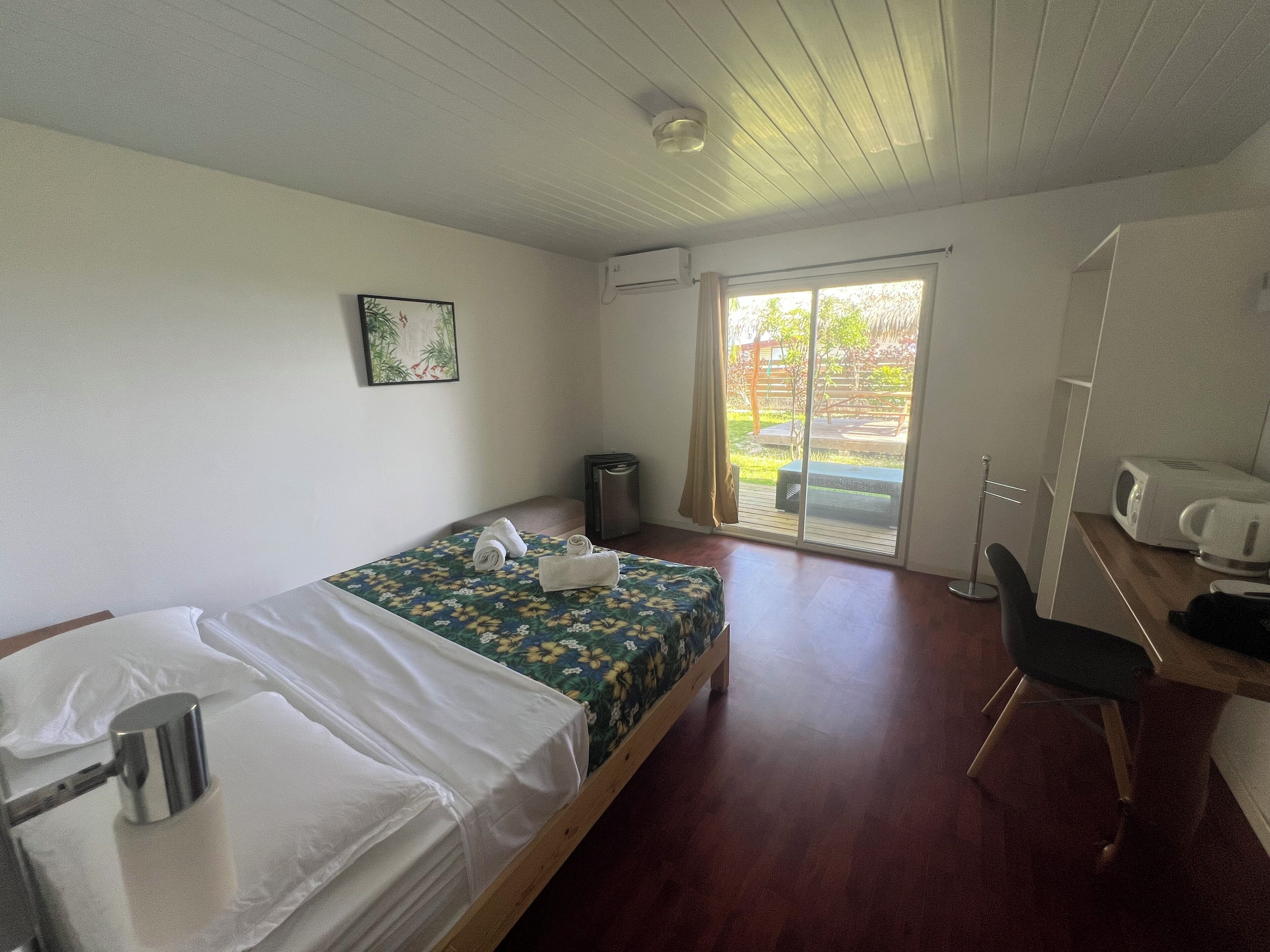 A single bed with a floral bedspread, a small bedside table with a lamp, a chair, and a view of a garden through a sliding door.