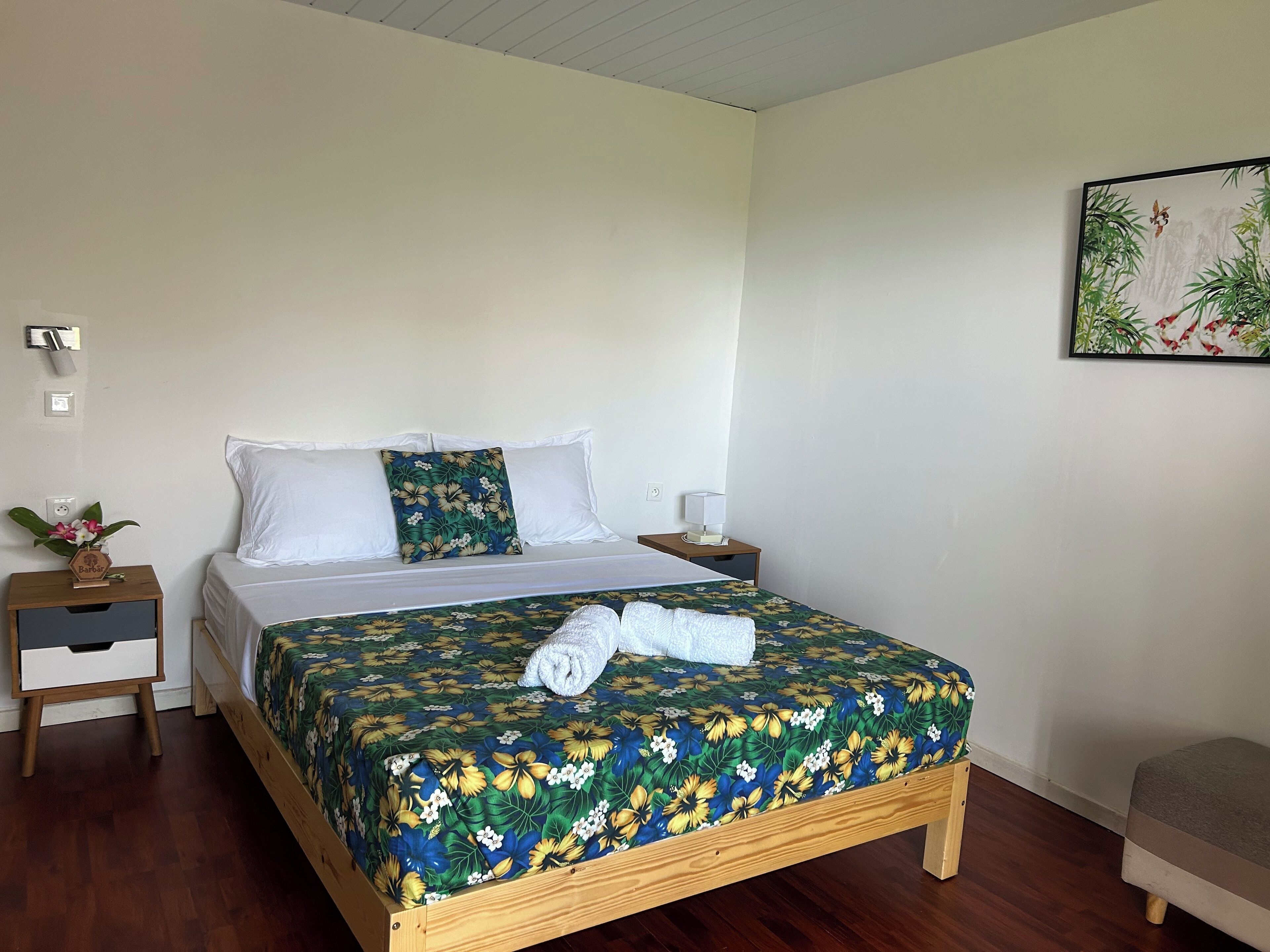 A neatly arranged bedroom with a wooden bed, white pillows, a floral bedspread, a bedside table with a lamp, and a small nightstand.