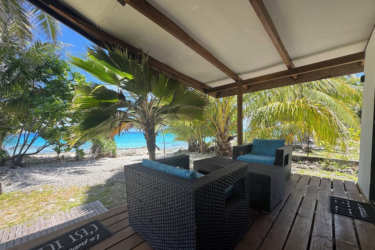 Uma área de estar coberta ao ar livre com móveis de vime, um deck de madeira e vista para a praia e o oceano.