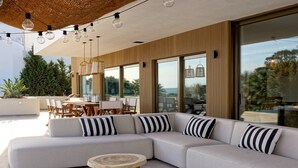 A modern outdoor seating area with a sectional sofa, round wooden tables, and large windows offering a view of the outdoors. - Luxury Beach Villa - Casa do Forte- Infinity pool. (Albufeira)