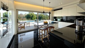 A modern kitchen with a large island, black countertops, and a view of a pool and greenery outside. - Luxury Beach Villa - Casa do Forte- Infinity pool. (Albufeira)