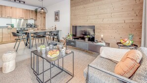 A modern living room with a sofa, coffee table, and TV. The room has wooden paneling on one wall and a kitchen area with a dining table and chairs.