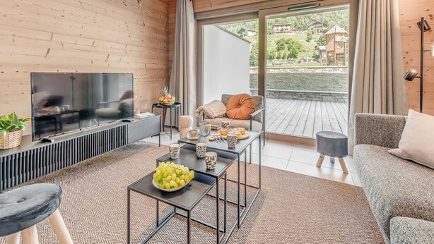A modern living room with a flat-screen TV, a coffee table with fruits, a sofa, and a balcony with curtains.