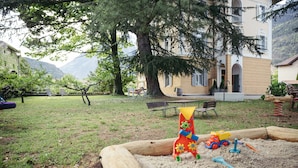 A sandy play area with toys, a wooden bench, and a building with a balcony in the background.