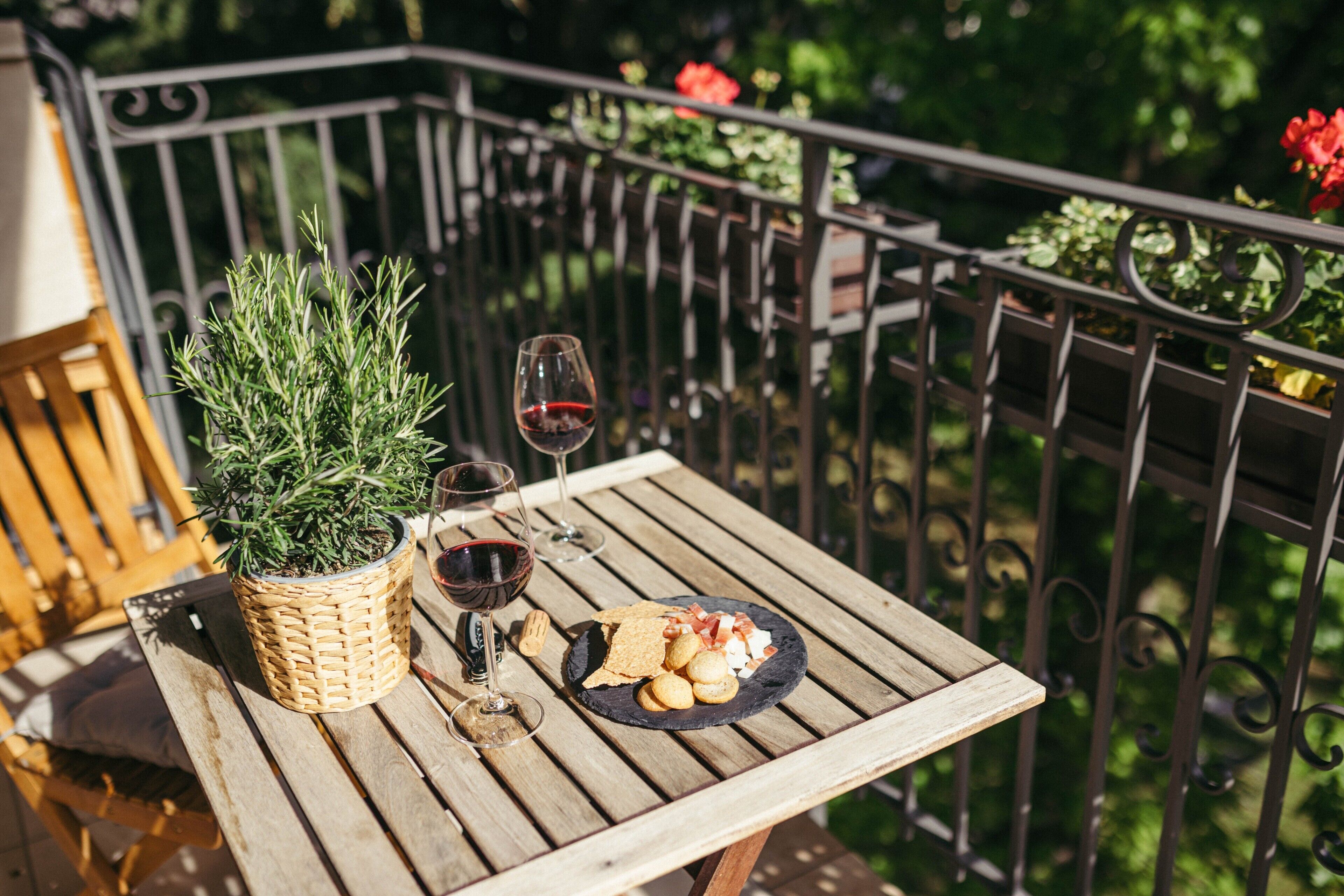 A wooden table with a potted plant, wine glasses, and a plate of snacks on a balcony.