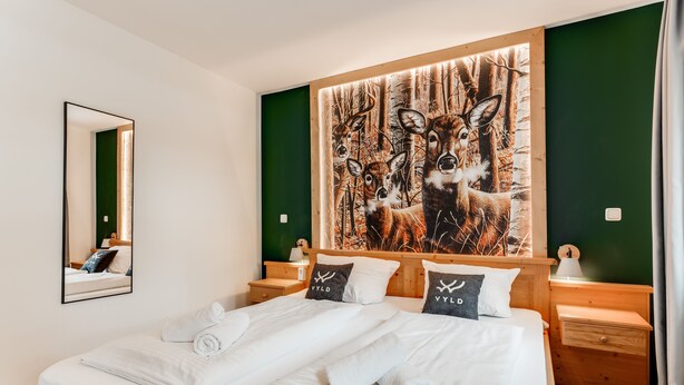 Ein ordentlich bezogenes Bett mit weißen Leinen und zwei Kissen, ein hölzernes Kopfteil, ein Nachttisch mit Lampe sowie ein großes Bild von einem Hirsch an der Wand.