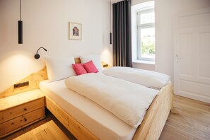 Un dormitorio con cama de madera, ropa de cama blanca, una mesita de noche, una lámpara de pared, una ventana con cortinas y una puerta.