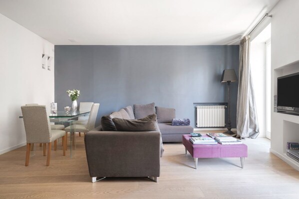 Un salon moderne avec un canapé gris, une table à manger en verre avec des chaises, un pouf rose et un téléviseur à écran plat fixé au mur.