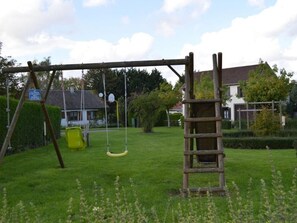 A wooden swing set in a grassy yard with a ladder, a yellow slide, and a house in the background.