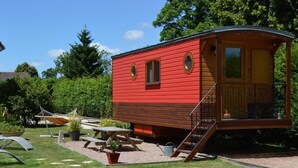 Una cabaña roja con balcón, rodeada de un área verde con mesas de picnic y plantas en macetas.
