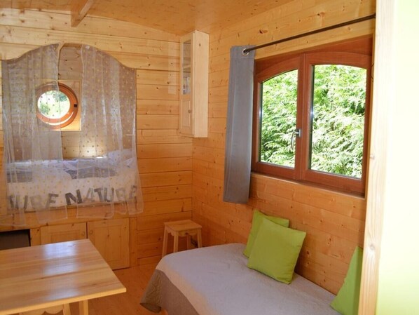 A wooden cabin room with a bed, a small table, a window with curtains, and a view of greenery outside.