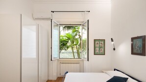 Un dormitorio con cama, ventana con vista a follaje, radiador y lámpara fijada a la pared.
