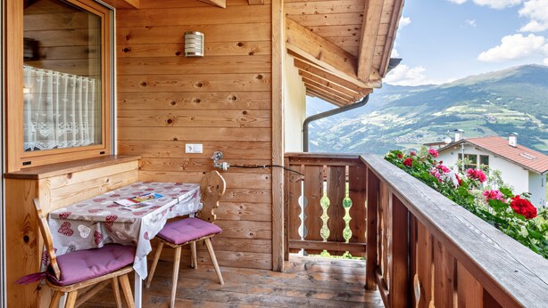 A wooden balcony with a table and chairs, a window with lace curtains, and a view of a mountain landscape.