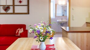 A wooden table with a white vase containing a floral arrangement, a red couch, and a kitchen area in the background.
