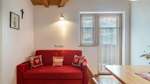 Een rode bank met gekleurde kussens, een houten tafel, een wandlamp, een raam met uitzicht en een lijst met een afbeelding aan de muur.