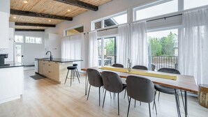 Moderni keittiö, jossa on puupalkkeja, ruokailutila pöytineen ja tuoleineen sekä suuret ikkunat, joissa on verhot.