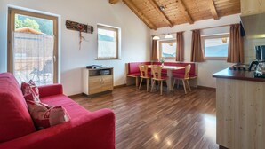 A cozy room with a red sofa, wooden ceiling beams, a dining area with a table and chairs, and large windows with curtains. - Apartment 'Solerhof Terrace' with Mountain View, Private Terrace and Wi-Fi (Laion)