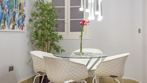 A dining area with a glass table, white chairs, a vase with pink flowers, a bamboo plant, and a colorful abstract painting on the wall.