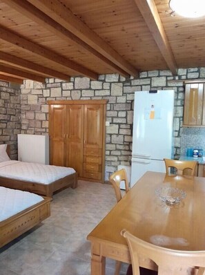 Una habitación con techo de madera, paredes de piedra, una cama, una mesa con sillas y un refrigerador.