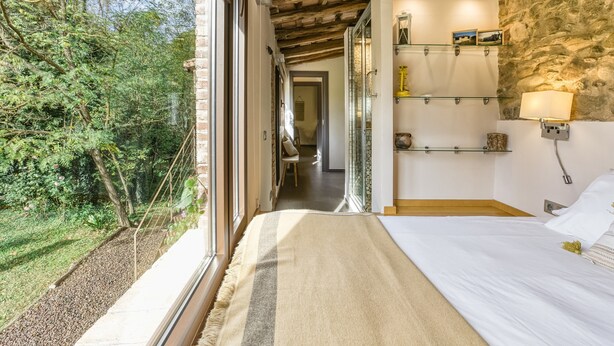A bedroom with a large window, a bed, a stone wall, and a shelf with items.