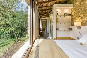 Un dormitorio con un ventanal grande, una cama, una pared de piedra y un estante con objetos.