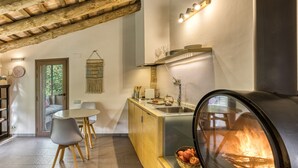 A modern kitchen with a wood-beamed ceiling, a round wood-burning stove, a dining area with a table and chairs, and a view of greenery through a door.