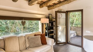 A room with a wooden ceiling, a sofa with cushions, a bookshelf, and a view of a balcony with a chair.